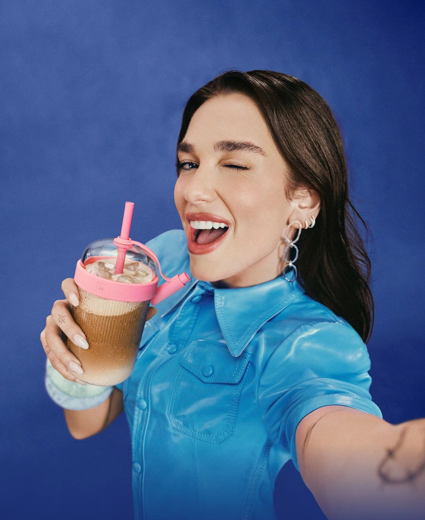 Dua Lipa holding a pink iced coffee drink against a bright blue background
